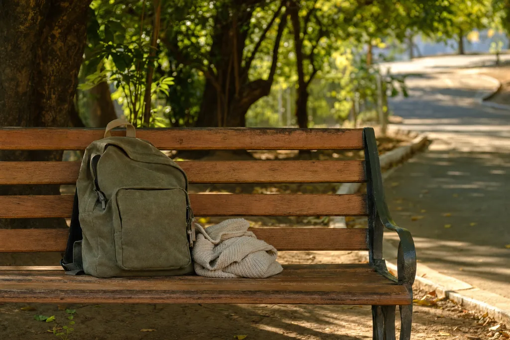 Banco de madeira em praça arborizada no fim da tarde com uma mochila apoiada em um dos lados e o outro lugar vazio, luz quente filtrada pelas folhas
